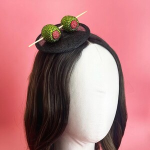 May include: A black fascinator with two green beaded olive decorations, each with a red centre, held together by a wooden cocktail stick. This headpiece is designed to be worn on the head, offering a fun and unique style.