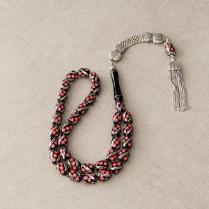 Könnte beinhalten: Eine Gebetskette mit schwarzen, ovalen Perlen, die mit roten kreisförmigen Mustern und weißen Akzenten verziert sind. Die Kette enthält eine glatte, schwarze zylindrische Perle, silberfarbene Metallzwischenstücke und eine silberfarbene Quaste.