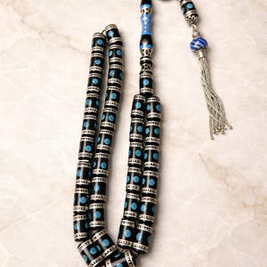 Peut inclure: Un chapelet orné de perles noires et argentées avec des accents turquoise. Les perles cylindriques présentent des bandes argentées et des motifs de points turquoise. Un gland argenté est suspendu à l'extrémité, et le haut présente un élément décoratif bleu et argenté.