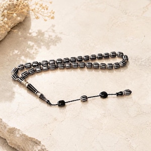 Peut inclure: Un chapelet composé de perles noires en forme de tonneau avec des rayures blanches verticales. Le cordon comprend une longue perle noire effilée avec des accents en métal argenté, suivie de trois perles plus petites et d'un embout décoratif.