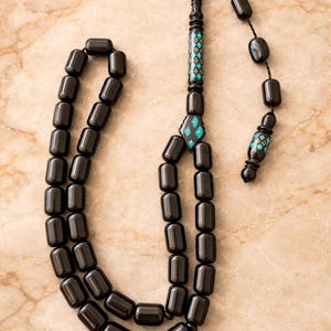 May include: A black prayer bead necklace with turquoise and black decorative elements. The beads are rectangular and strung together. Geometric patterns decorate the top and bottom sections of the necklace. The necklace is made of small beads.