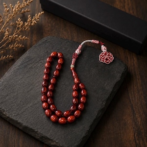 Peut inclure: Un chapelet de prière rouge foncé avec des perles ovales, posé sur une pierre grise foncée. Le chapelet a un pompon décoratif et un cordon argenté et rouge. Une boîte noire est en arrière-plan.