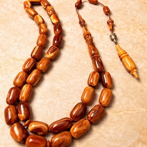 Peut inclure: Un chapelet avec des perles ovales dans les tons marron et beige. Les perles sont enfilées avec un cordon assorti, et l'ensemble comprend un gland décoratif. Le fond est une surface texturée brun clair.