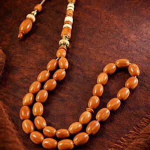 Può includere: Una collana di grani di preghiera con perline di forma ovale nei toni del marrone. Le perline sono infilate con un nappa decorativa e accenti bianchi e dorati. Lo sfondo è una superficie strutturata marrone scuro.