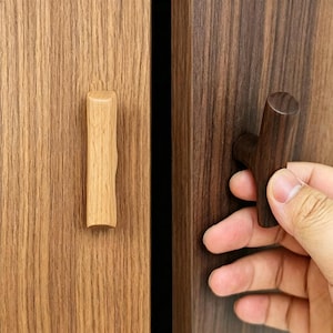 Può includere: Maniglie per ante di armadi in legno. Una è di colore chiaro, di forma rettangolare verticale con un bordo strutturato. L'altra è una maniglia a forma di T, marrone scuro. Le ante dell'armadio hanno un motivo a venature del legno.