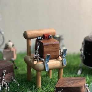 Puede incluir: Una figura de robot de madera con un cuerpo cuadrado marrón oscuro, brazos y piernas de metal plateado y una pequeña cámara roja en la parte superior. El robot está sentado en una silla de madera en miniatura. Otras figuras de robots están en el fondo.