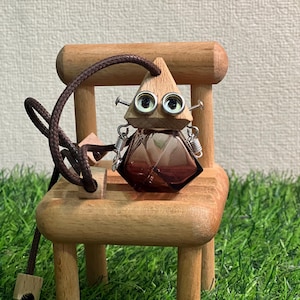 Puede incluir: Un adorno caprichoso de robot de madera con un cuerpo de vidrio marrón y una cabeza triangular de madera. El robot tiene brazos de resorte de metal y ojos de tornillo. Se sienta en una silla de madera en miniatura con un cordón marrón.