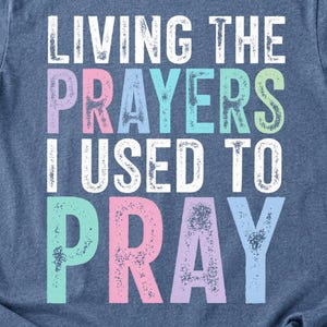 Peut inclure: Un t-shirt bleu chiné à manches courtes avec le texte blanc et pastel "LIVING THE PRAYERS I USED TO PRAY" imprimé sur le devant. Le t-shirt présente un col rond, une coupe standard avec des manches retroussées et un motif imprimé usé.
