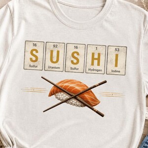 Peut inclure: Un t-shirt couleur crème arborant un graphique du mot SUSHI épelé avec des éléments du tableau périodique : Soufre, Uranium, Soufre, Hydrogène et Iode. Sous le texte se trouve une illustration d'un nigiri au saumon avec des baguettes croisées.