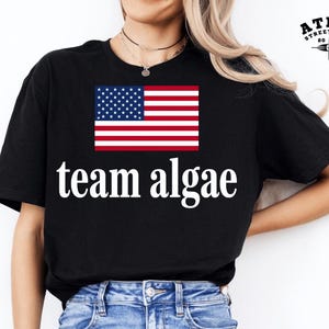 Peut inclure: Un t-shirt noir à manches courtes arborant un graphique du drapeau des États-Unis placé au-dessus du texte blanc "team algae" en police serif. Le t-shirt est associé à un jean bleu clair présentant des déchirures sur le haut des cuisses.