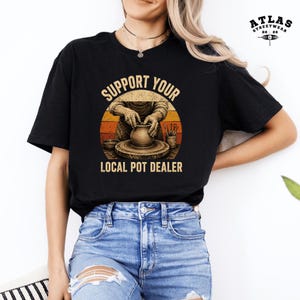 Peut inclure: Un t-shirt noir à manches courtes avec un motif représentant une personne façonnant de l'argile sur un tour de potier. Le design inclut un fond circulaire aux couleurs de coucher de soleil et le texte "SUPPORT YOUR LOCAL POT DEALER".