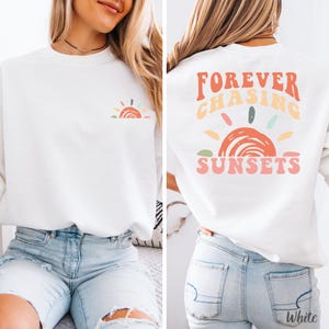 May include: White sweatshirt with a colorful sunset graphic on the front left chest and back. The back of the sweatshirt features the text "FOREVER CHASING SUNSETS" in a retro font. The design includes shades of orange, pink, yellow, and green.