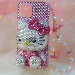 May include: Pink rhinestone-covered phone case featuring a Hello Kitty design. The case has a bow, eyes, and whiskers made of rhinestones. A pink strap with decorative beads and charms is attached to the case.