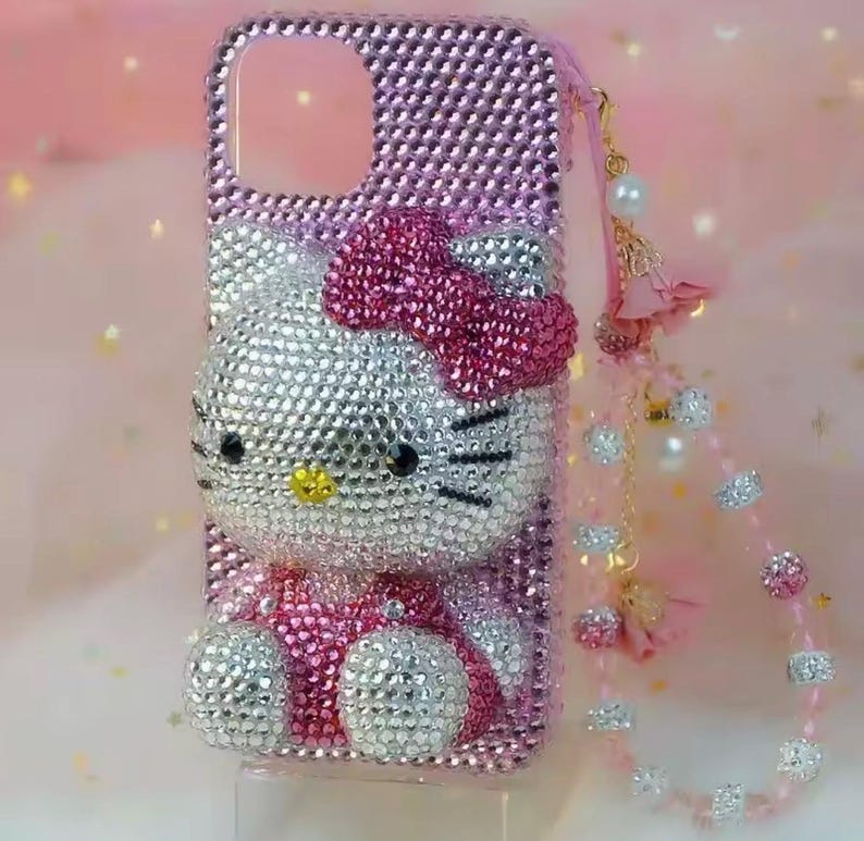 May include: Pink rhinestone-covered phone case featuring a Hello Kitty design. The case has a bow, eyes, and whiskers made of rhinestones. A pink strap with decorative beads and charms is attached to the case.