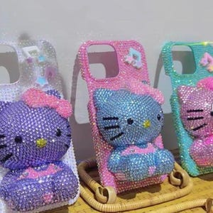 Puede incluir: Tres fundas de tel&eacute;fono adornadas con pedrer&iacute;a con un dise&ntilde;o de Hello Kitty. Las fundas est&aacute;n incrustadas con pedrer&iacute;a en varios colores, incluyendo morado, rosa y azul. Cada funda tiene un esquema de color y dise&ntilde;o diferente, con un lazo en la parte superior.
