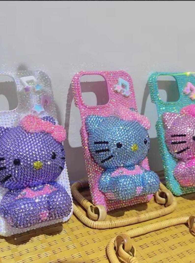 Puede incluir: Tres fundas de tel&eacute;fono adornadas con pedrer&iacute;a con un dise&ntilde;o de Hello Kitty. Las fundas est&aacute;n incrustadas con pedrer&iacute;a en varios colores, incluyendo morado, rosa y azul. Cada funda tiene un esquema de color y dise&ntilde;o diferente, con un lazo en la parte superior.