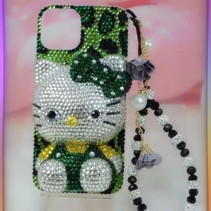 May include: A bedazzled phone case featuring a Hello Kitty design. The case is covered in sparkling rhinestones, with a green and black leopard print pattern on the top. A charm with pearls and beads hangs from the case.