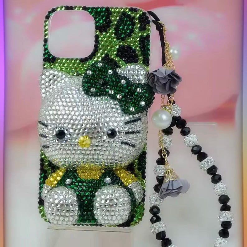 May include: A bedazzled phone case featuring a Hello Kitty design. The case is covered in sparkling rhinestones, with a green and black leopard print pattern on the top. A charm with pearls and beads hangs from the case.