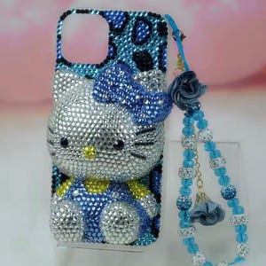 May include: A bedazzled phone case featuring a Hello Kitty design. The case is covered in silver, blue, and yellow rhinestones. A matching blue beaded wrist strap with a flower charm is attached.
