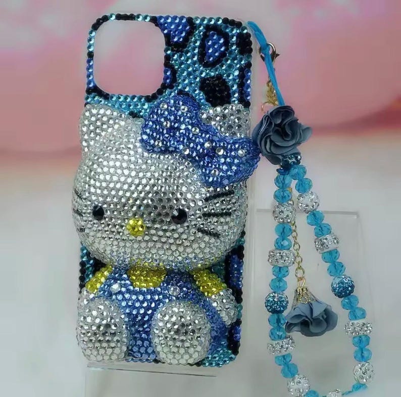 May include: A bedazzled phone case featuring a Hello Kitty design. The case is covered in silver, blue, and yellow rhinestones. A matching blue beaded wrist strap with a flower charm is attached.