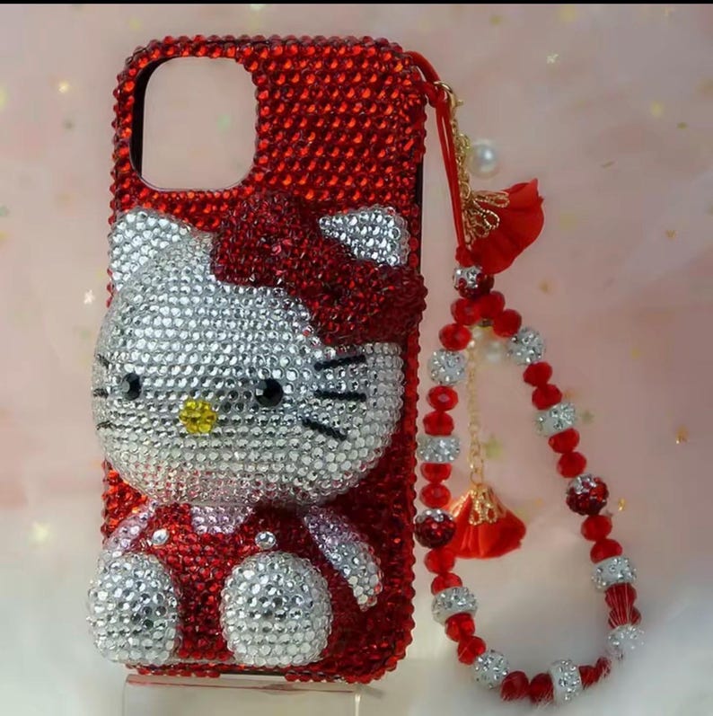 May include: Red rhinestone-covered phone case featuring a Hello Kitty design. The case has a matching red bow and a beaded wrist strap with red and silver accents. The phone case is designed to fit a specific phone model.