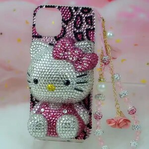 May include: A bedazzled phone case featuring a Hello Kitty design. The case is covered in silver and pink rhinestones, with a pink bow and a leopard print accent. A charm with pearls and pink flowers hangs from the case.