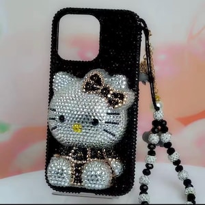 May include: Black phone case with a sparkling Hello Kitty design. The case is covered in small, shiny crystals, with a bow and accents in brown and gold. A beaded strap with black and clear beads is attached.