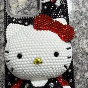 Puede incluir: Una funda de tel&eacute;fono adornada con pedrer&iacute;a, con un dise&ntilde;o de Hello Kitty blanco con un lazo rojo y detalles en negro. La funda est&aacute; cubierta de peque&ntilde;os cristales brillantes, creando un efecto texturizado. El dise&ntilde;o incluye detalles en blanco y negro.