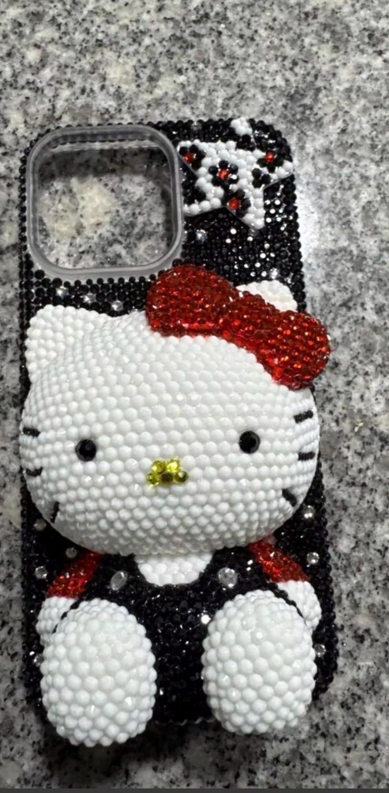 Puede incluir: Una funda de tel&eacute;fono adornada con pedrer&iacute;a, con un dise&ntilde;o de Hello Kitty blanco con un lazo rojo y detalles en negro. La funda est&aacute; cubierta de peque&ntilde;os cristales brillantes, creando un efecto texturizado. El dise&ntilde;o incluye detalles en blanco y negro.