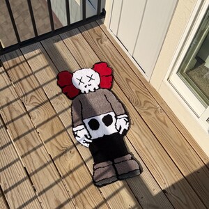 May include: A handmade rug in the shape of a cartoon character with a skull-like face and crossed-out eyes. The rug features a gray body, black pants, and brown shoes, with red ear-like shapes on the head. The rug is placed on a wooden deck.