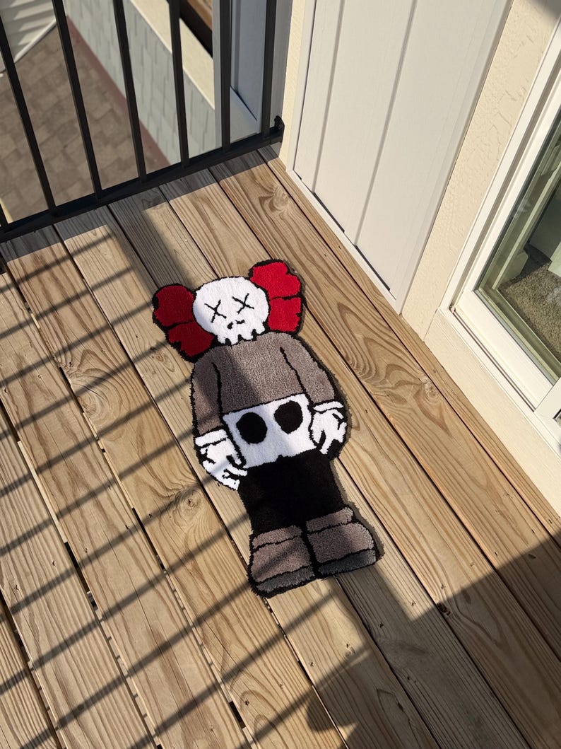 May include: A handmade rug in the shape of a cartoon character with a skull-like face and crossed-out eyes. The rug features a gray body, black pants, and brown shoes, with red ear-like shapes on the head. The rug is placed on a wooden deck.