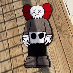 May include: A handmade rug in the shape of a cartoon character. The rug features a white skull-like head with black cross-shaped eyes, red ears, a gray body, and black and gray legs. The rug is placed on a wooden surface.