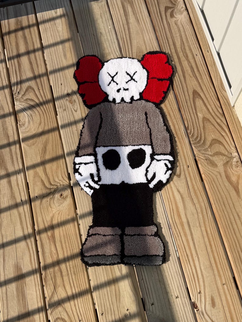 May include: A handmade rug in the shape of a cartoon character. The rug features a white skull-like head with black cross-shaped eyes, red ears, a gray body, and black and gray legs. The rug is placed on a wooden surface.