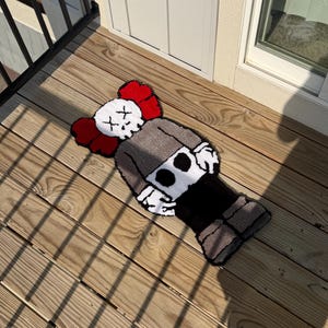 May include: A handmade rug featuring a cartoon character design with a white skull-like face, red ears, and a gray body. The rug is placed on a wooden deck, showcasing its unique design and texture. The rug's dimensions are not visible.