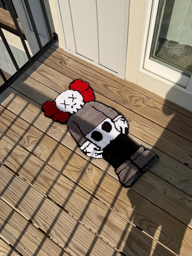 May include: A handmade rug featuring a cartoon character design with a white skull-like face, red ears, and a gray body. The rug is placed on a wooden deck, showcasing its unique design and texture. The rug's dimensions are not visible.
