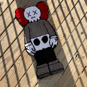 May include: A handmade rug in the shape of a cartoon character with a skull-like face and crossed-out eyes. The rug features a gray body, black pants, and red ear-shaped accents. The rug is approximately 30 inches long.