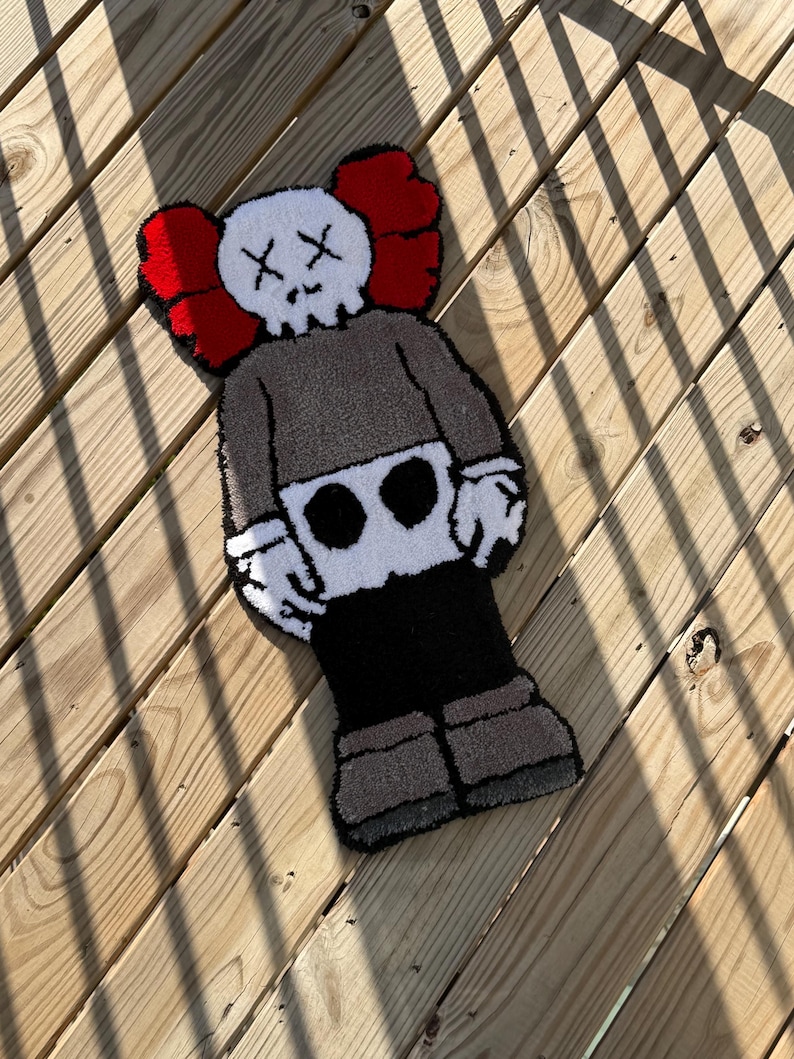 May include: A handmade rug in the shape of a cartoon character with a skull-like face and crossed-out eyes. The rug features a gray body, black pants, and red ear-shaped accents. The rug is approximately 30 inches long.