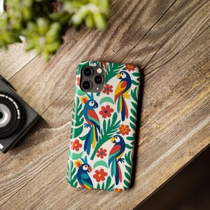 Peut inclure: Une coque de téléphone ornée d'un motif coloré de perroquets et de fleurs. La coque présente un fond blanc avec des illustrations de perroquets bleus, rouges et jaunes, de fleurs rouges et de feuilles vertes. Posée sur une surface en bois.
