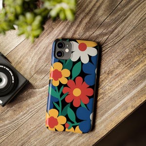 Peut inclure: Coque de téléphone à motifs floraux avec des fleurs rouges, jaunes, bleues et blanches sur fond noir. L'étui brillant est posé sur une surface en bois.