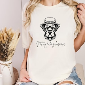 Könnte beinhalten: Ein cremefarbenes T-Shirt mit einer schwarzen Grafik eines Hundes, der einen Hut und eine Brille trägt. Unter dem Hund steht der Text: "Not my fucking business." Das T-Shirt hat kurze Ärmel und einen Rundhalsausschnitt für einen schlichten Look.