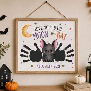 Puede incluir: Una impresi&oacute;n de arte de pared enmarcada con una ilustraci&oacute;n central de un peque&ntilde;o murci&eacute;lago gris con orejas rosas, flanqueado por dos huellas de manos negras. El texto dice LOVE YOU TO THE MOON AND BAT y HALLOWEEN 2026, con una luna creciente.