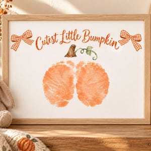 Puede incluir: Un marco de madera rectangular que contiene una impresión blanca con el texto naranja Cutest Little Bumpkin. El diseño presenta dos huellas de pies de bebé naranjas con forma de calabaza, acentuadas por dos lazos de cuadros naranjas.