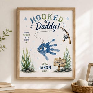 Puede incluir: Un estampado enmarcado con las palabras "Hooked on Daddy!" y un diseño de pez con huellas de manos. El estampado incluye el texto "You're the best catch ever!" y "Happy Father's Day!" en estilo acuarela. El estampado está personalizado con el nombre "Jaxon" y el año 2026.