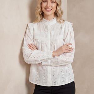 Puede incluir: Blusa blanca de ojal con cuello alto con volantes y mangas largas. La blusa presenta una parte delantera abotonada y detalles intrincados calados. La modelo lleva pantalones negros.