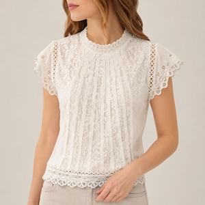 May include: A white short-sleeved blouse featuring intricate floral lace detailing, a high neckline with a scalloped edge, vertical pleated panels across the front, and delicate eyelet trim along the cap sleeves and the bottom hemline.