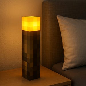 Peut inclure: Lampe en forme de torche Minecraft, avec un dessus éclairé jaune et orange et une base pixelisée marron foncé. La lampe est posée sur une table de chevet en bois, à côté d'un lit avec un oreiller.