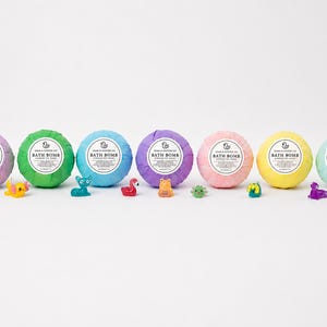 May include: Seven spherical bath bombs in purple, green, blue, lavender, pink, yellow, and light blue, each wrapped in paper with a white label reading "SOAK & SOOTHER CO BATH BOMB SURPRISE TOY INSIDE". Seven small plastic animal figurines are placed in front.