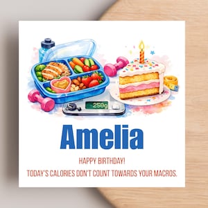 May include: A square greeting card featuring a watercolour illustration of a blue bento box with food, a slice of birthday cake with a candle, pink dumbbells, and a digital scale reading 250g. Text reads: Amelia, HAPPY BIRTHDAY! TODAY'S CALORIES DON'T COUNT TOWARDS YOUR MACROS.