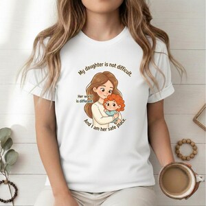 Puede incluir: Camiseta blanca con un gráfico de una madre y un niño y el texto: "Mi hija no es difícil. Su mundo es diferente. Y yo soy su lugar seguro."