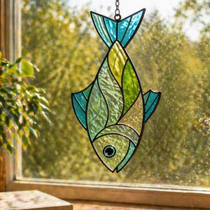 Könnte beinhalten: Ein Glasfenster-Fisch-Suncatcher mit schwarzer Umrandung, der vor einem Fenster hängt. Der Fisch hat Grün-, Gelb- und Blautöne. Der Suncatcher wird von einer Kette gehalten. Bücher und Glasstücke liegen auf der Holzoberfläche darunter.